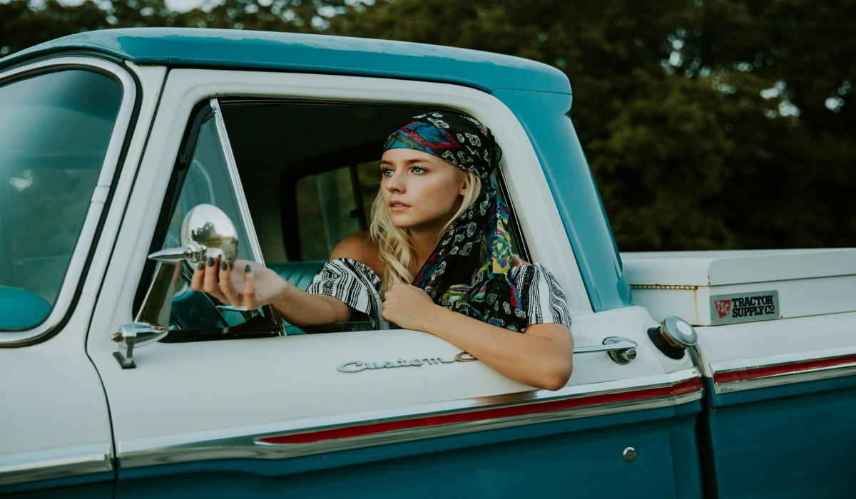Vintage Journey: Young Blonde Woman Adjusting Side Mirror in a Classic 2-Door Pickup Truck